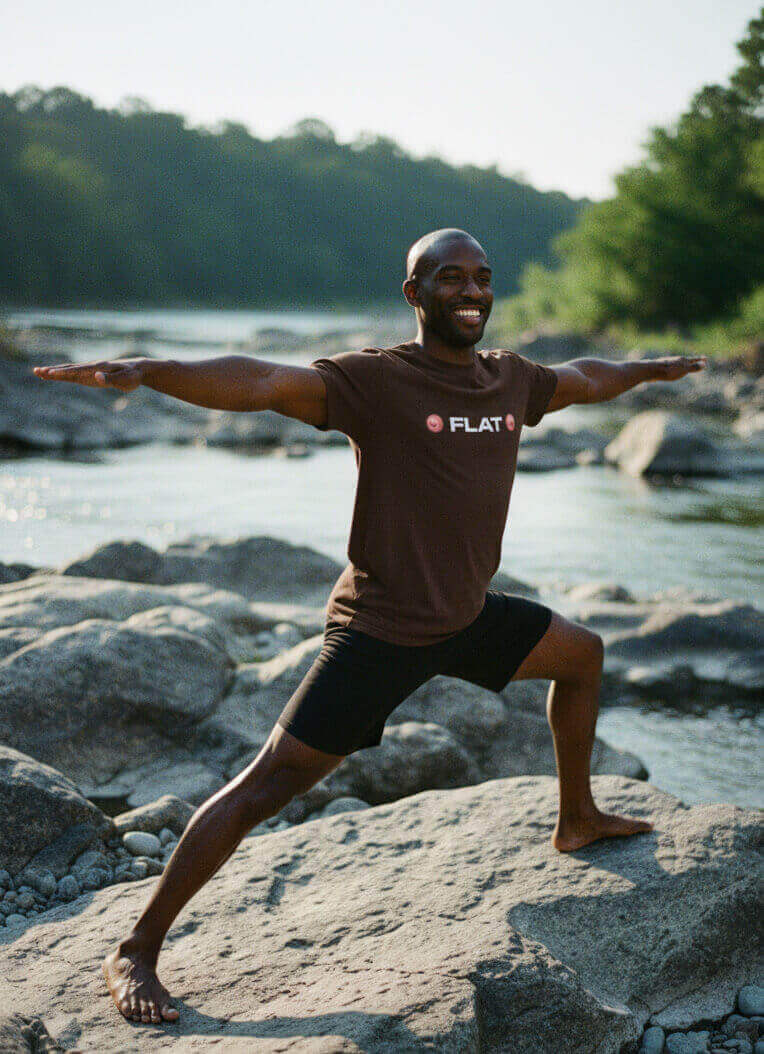 Man smiling while doing a yoga pose by a river, wearing the Espresso Brown Sovereign Emblem 'FLAT' t-shirt.
