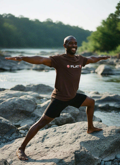 Man smiling while doing a yoga pose by a river, wearing the Espresso Brown Sovereign Emblem 'FLAT' t-shirt.
