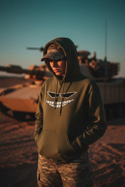 Female model in a military green Sovereign Grin hoodie and camo gear, standing in the desert with a tank in the background.