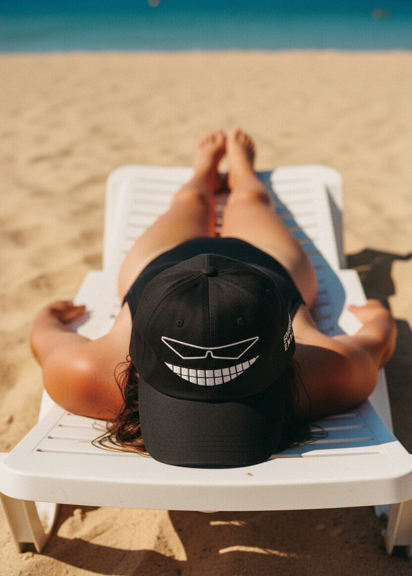 Woman relaxing on a beach lounger, wearing the black Sovereign Emblem "Sovereign Grin" dad cap.